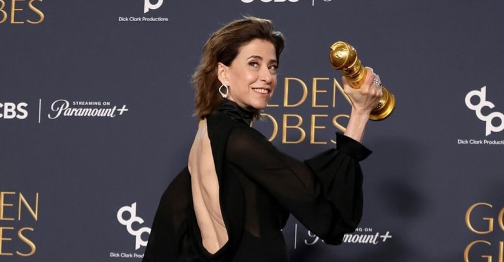 Foto mostra a Fernanda Torres segurnado o globo de ouro durante a premiação. A Fernanda está sorrindo de costas, com um vestido preto decotado nas costas e o rosto virado para o lado.