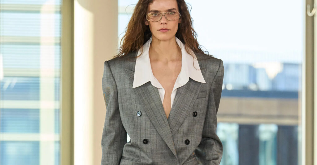 Imagem mostra uma modelo com óculos de grau, camisa e blaser cinza. Vestida em um estilo conserdor, que é uma das tendencias de destaque nas últimas semanas de moda.