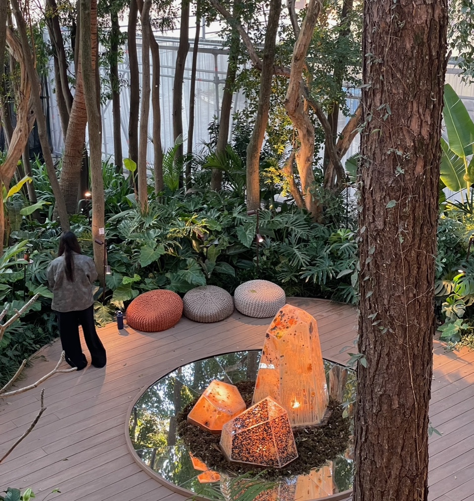 Exposição casa cor 2025. Foto mostra um ambiente externo com grandes pedras de cristais ao centro de um tablado circular de madeira, puffs e vegetação alta ao fundo.