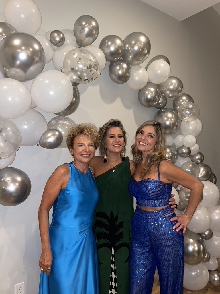 Foto da Silvia em aniversário com suas amigas. A Silvia usa vestido longo verde mais simples, combinando com a ocasião da festa.