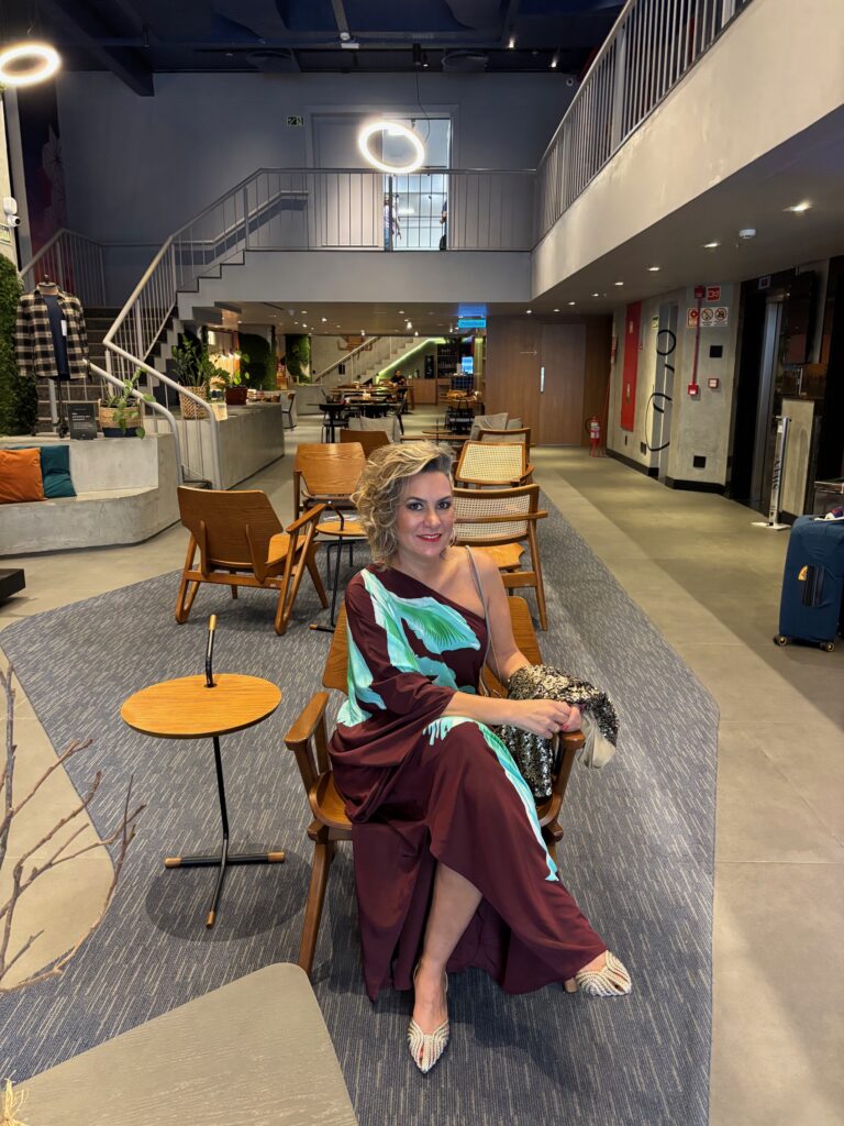 Foto da Silvia sentada em uma poltrona em evento. A Silvia usa um vestido longo marrom com uma estampa verde água de folhas. Também está com um sapato de salto mais baixo. O ambiente parece refinado e descontraído.