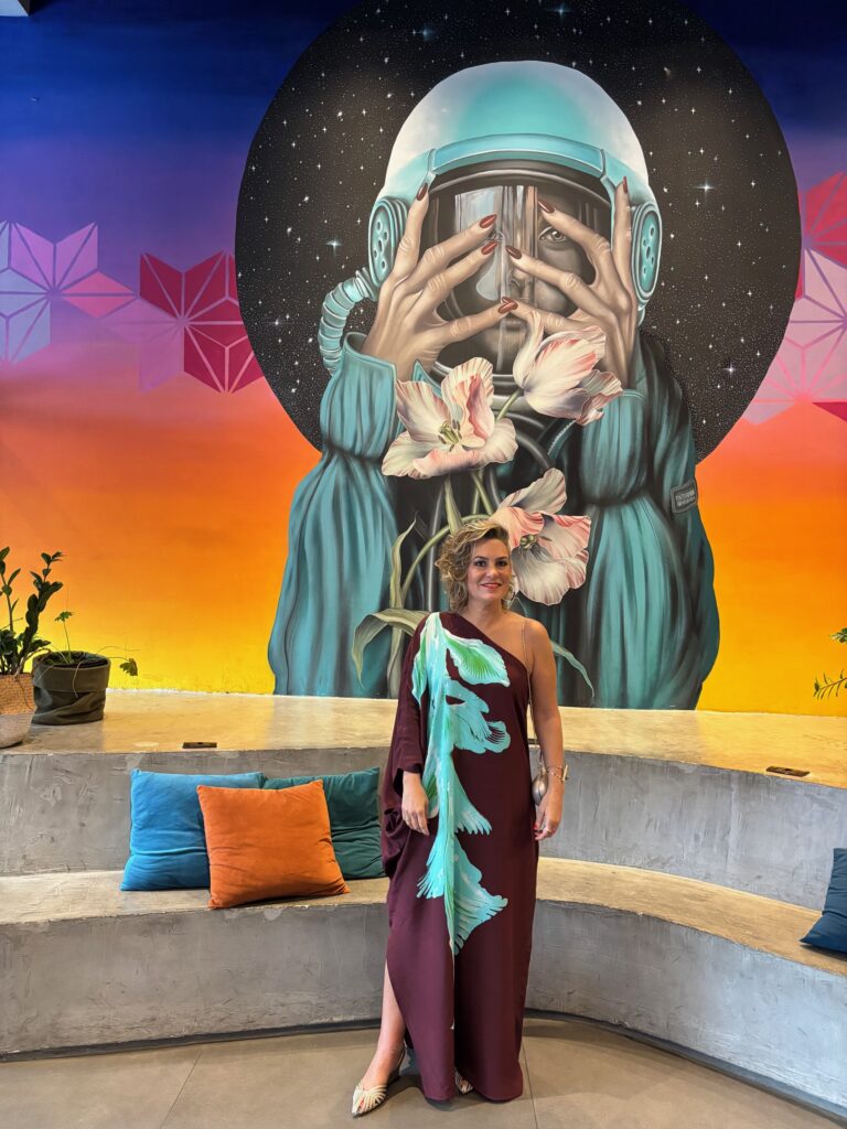 Foto da Silvia em pé com o mesmo vestido marrom. Ao fundo há uma pintura na parede com cores destacando.