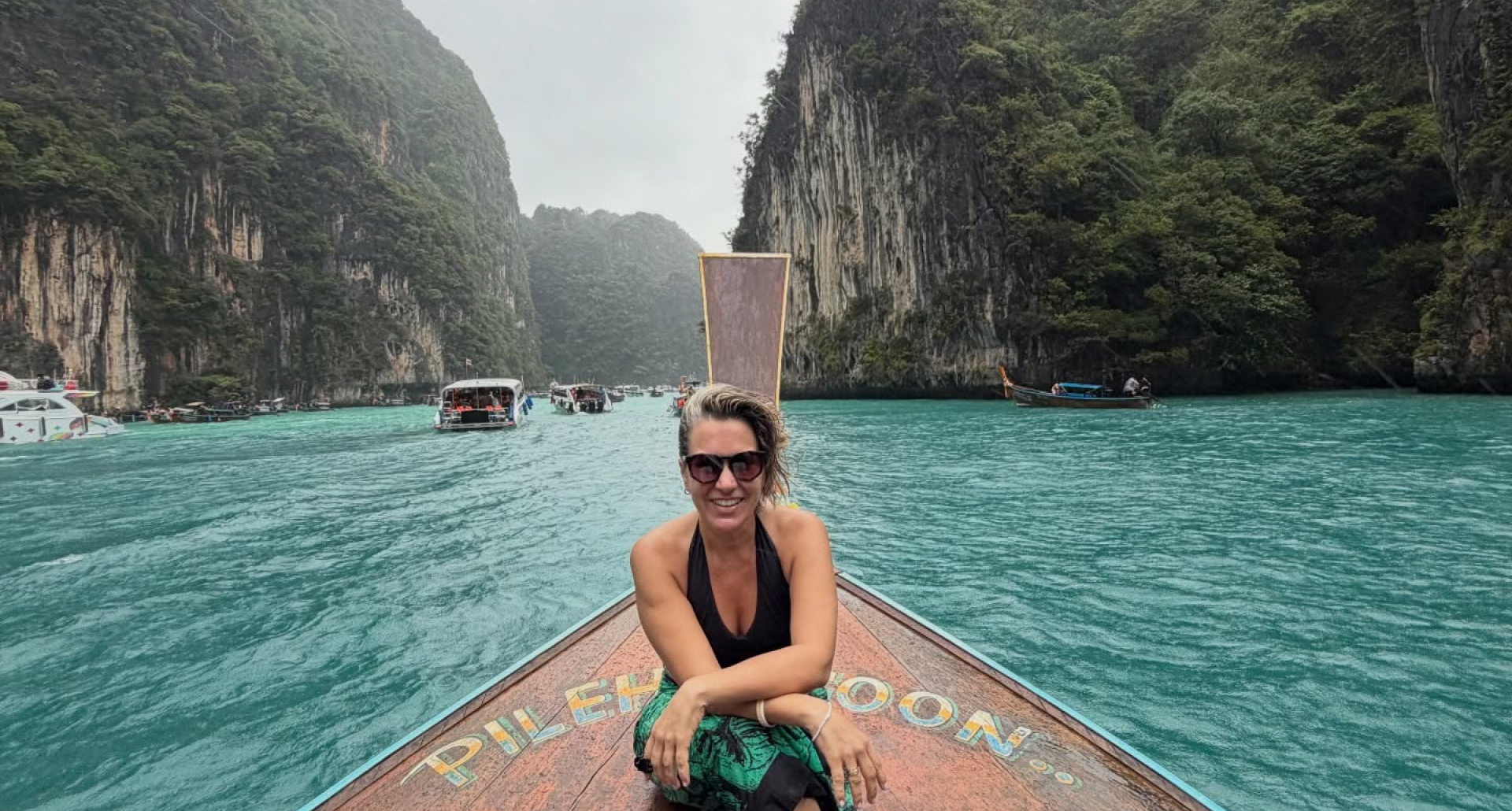 Foto da Silvia Scigliano na Tailandia. Na foto, ela está sentada na ponta de um barco sobre águas bem azuis. Ao fundo vê-se uma paisagem com morros verdes.