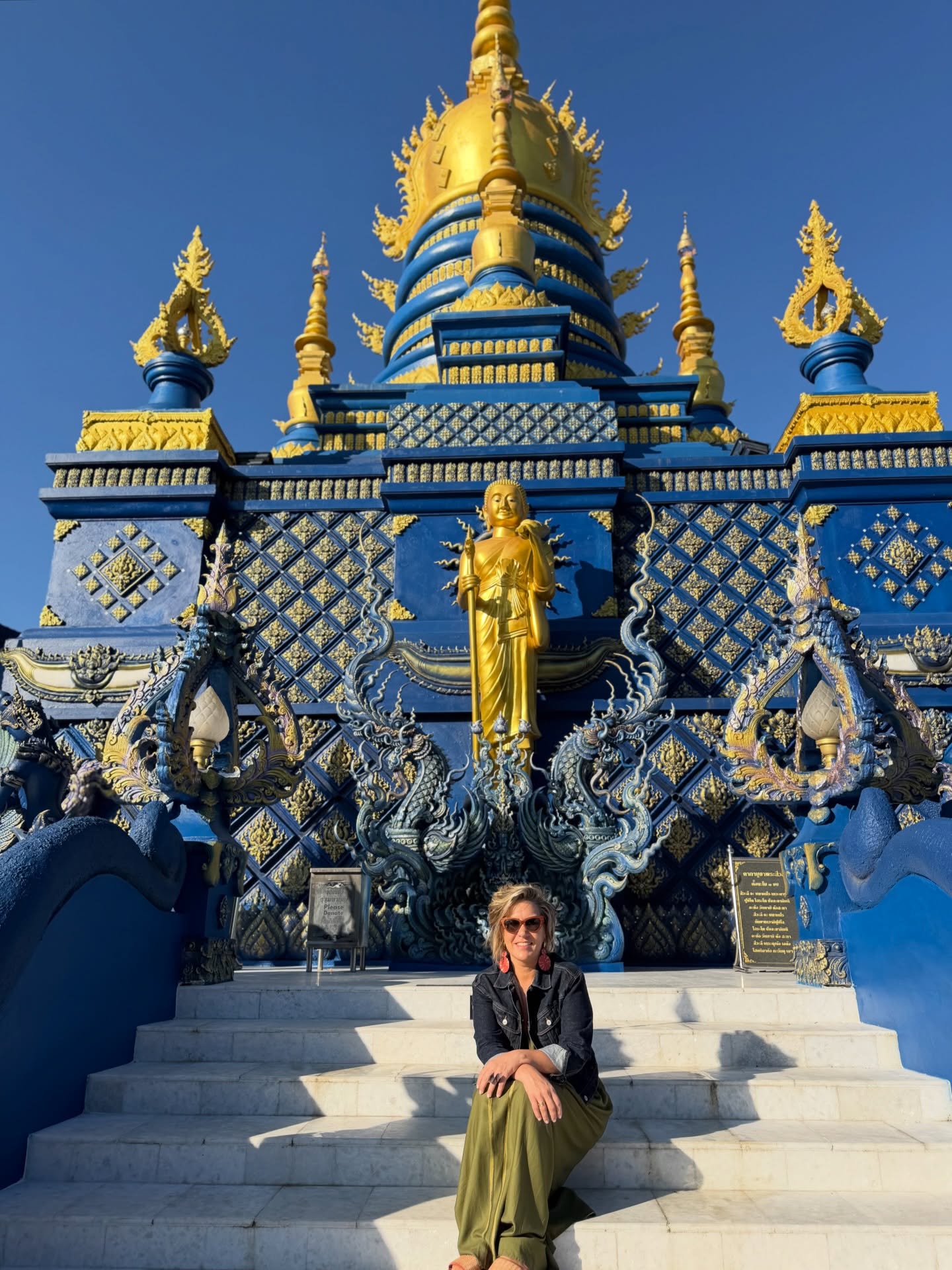Foto da Silvia sentada em escada na frente do templo azul da Tailândia, uma inspiração para a moda que combina riquesa de detalhes dourados.