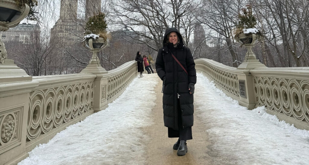 Foto da Silvia Scigliano vestida com botas e sobretudo com capuz em uma ponte com neve em Nova York