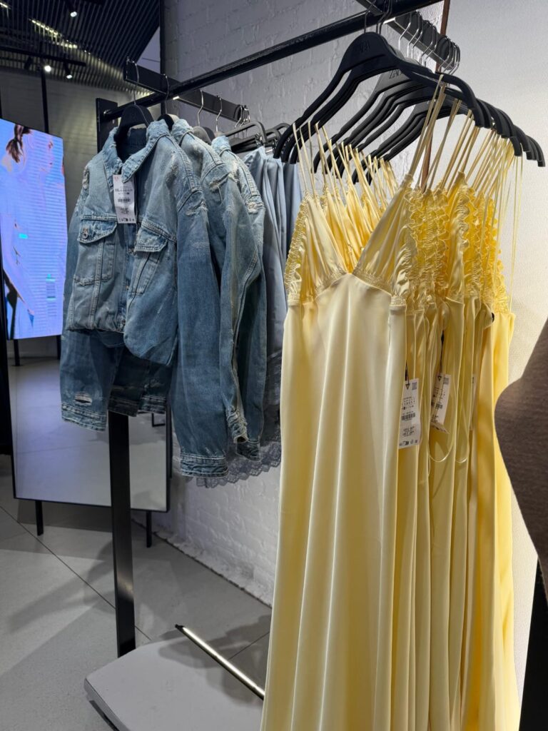 Foto de arara de loja com vestidos de cetim amarelo manteiga ao lado de jaqueta jeans.