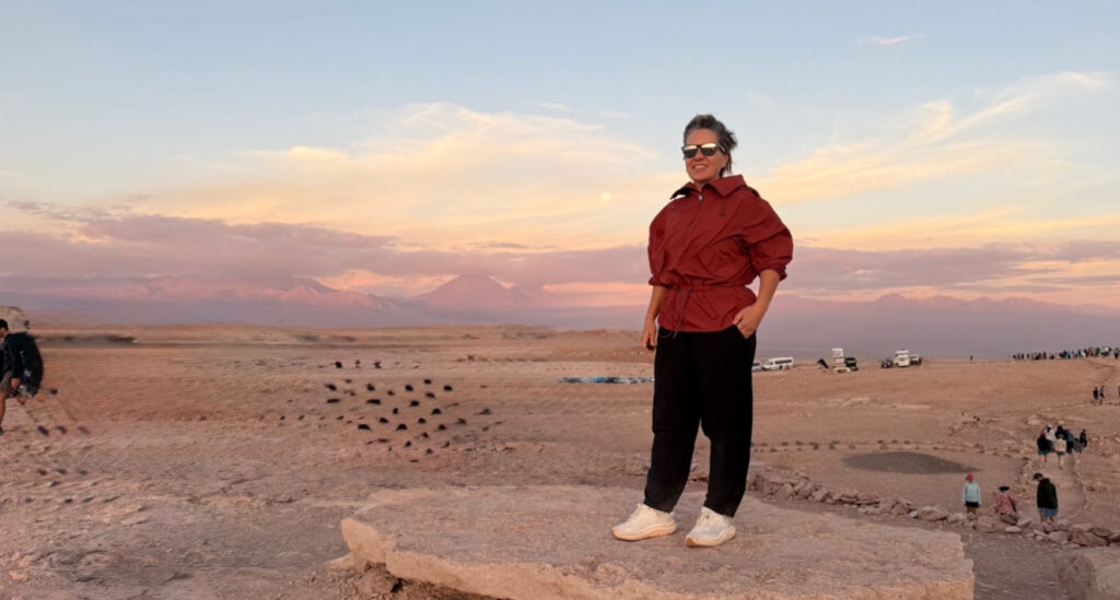 Foto de Silvia Sicliano, mostrando como se vestir no deserto. Na foto, Silvia usa óculos de sol, blusa vermelha estilo moletom e calça preta confortável, além de tênis.