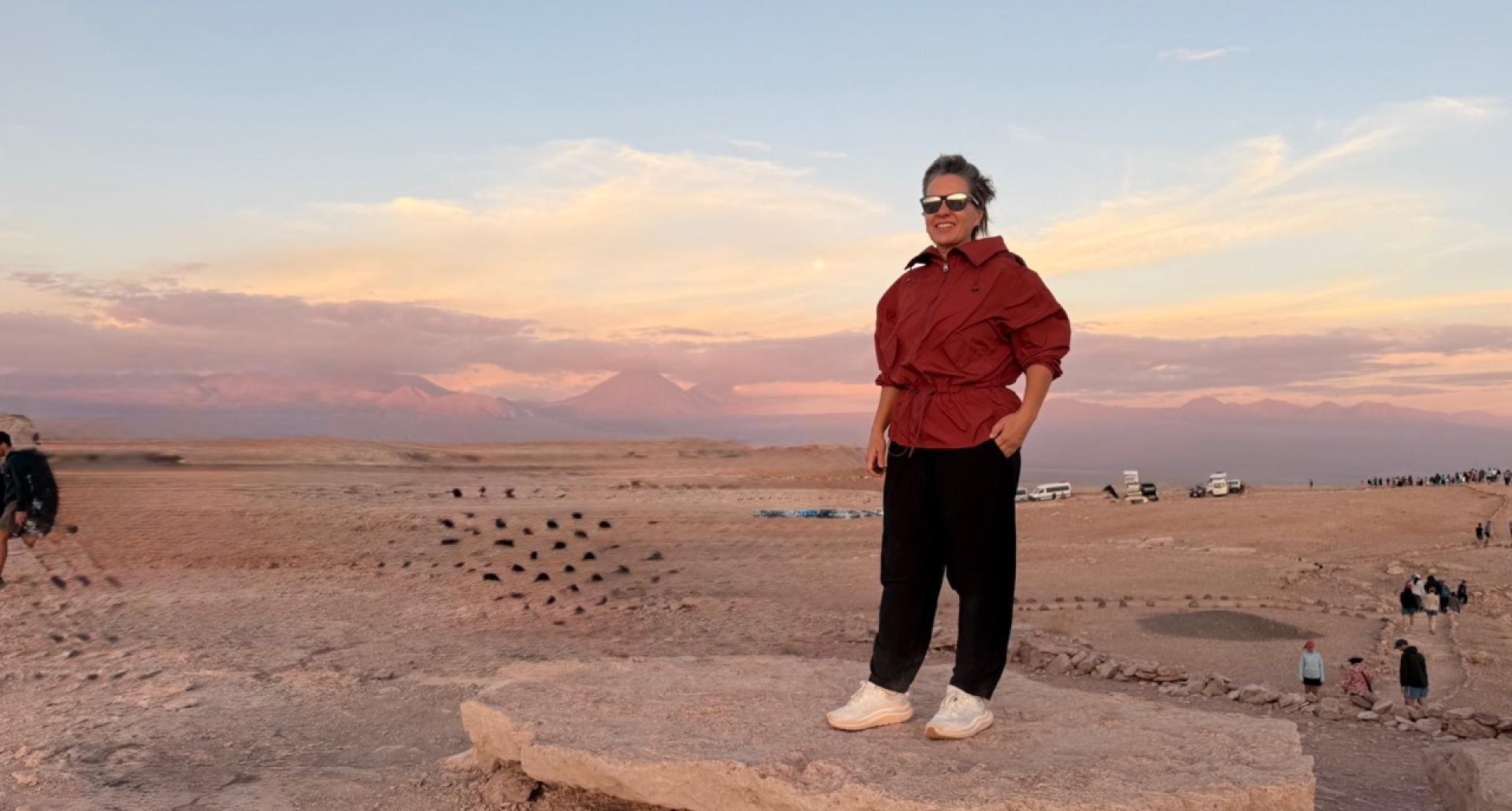 Foto de Silvia Sicliano, mostrando como se vestir no deserto. Na foto, Silvia usa óculos de sol, blusa vermelha estilo moletom e calça preta confortável, além de tênis.