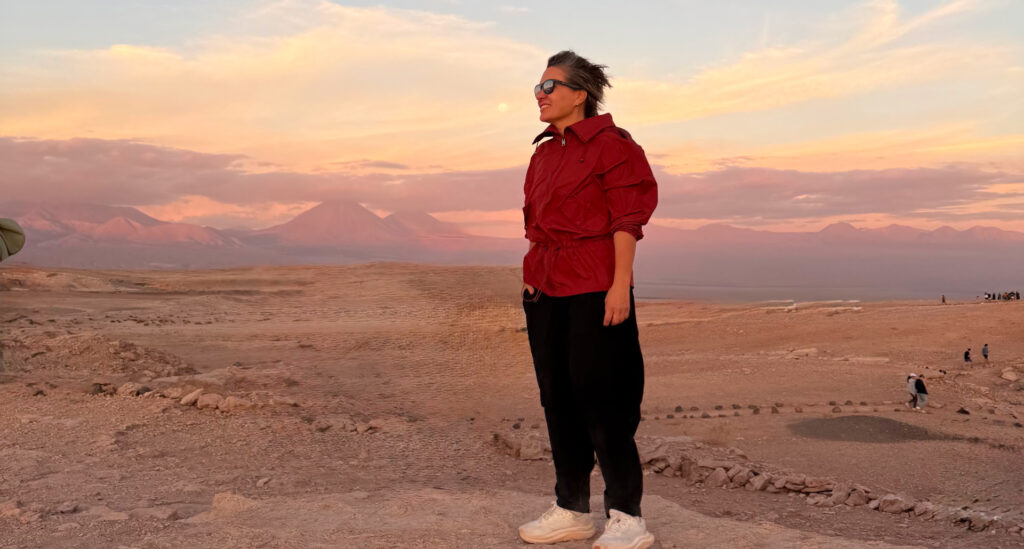 Foto de Silvia Sicliano, mostrando como se vestir no deserto. Na foto, Silvia está no deserto do Atacama e usa óculos de sol, blusa vermelha estilo moletom e calça preta confortável, além de tênis.