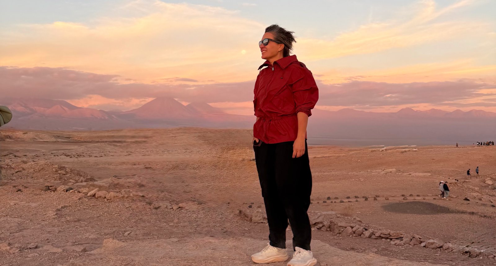 Foto de Silvia Sicliano, mostrando como se vestir no deserto. Na foto, Silvia está no deserto do Atacama e usa óculos de sol, blusa vermelha estilo moletom e calça preta confortável, além de tênis.